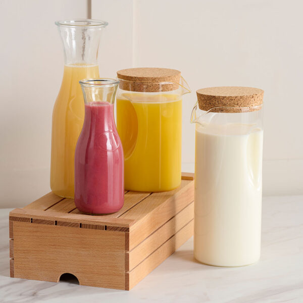 Clear Comas Buffet Jugs and Bottles with brown lids, displayed on a Comas Wooden Box and filled with orange juice, milk, and raspberry juice