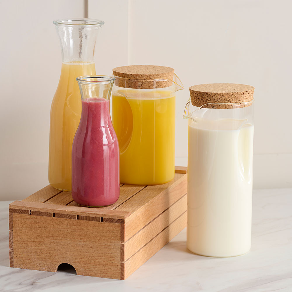 Clear Comas Buffet Jugs and Bottles with brown lids, displayed on a Comas Wooden Box and filled with orange juice, milk, and raspberry juice