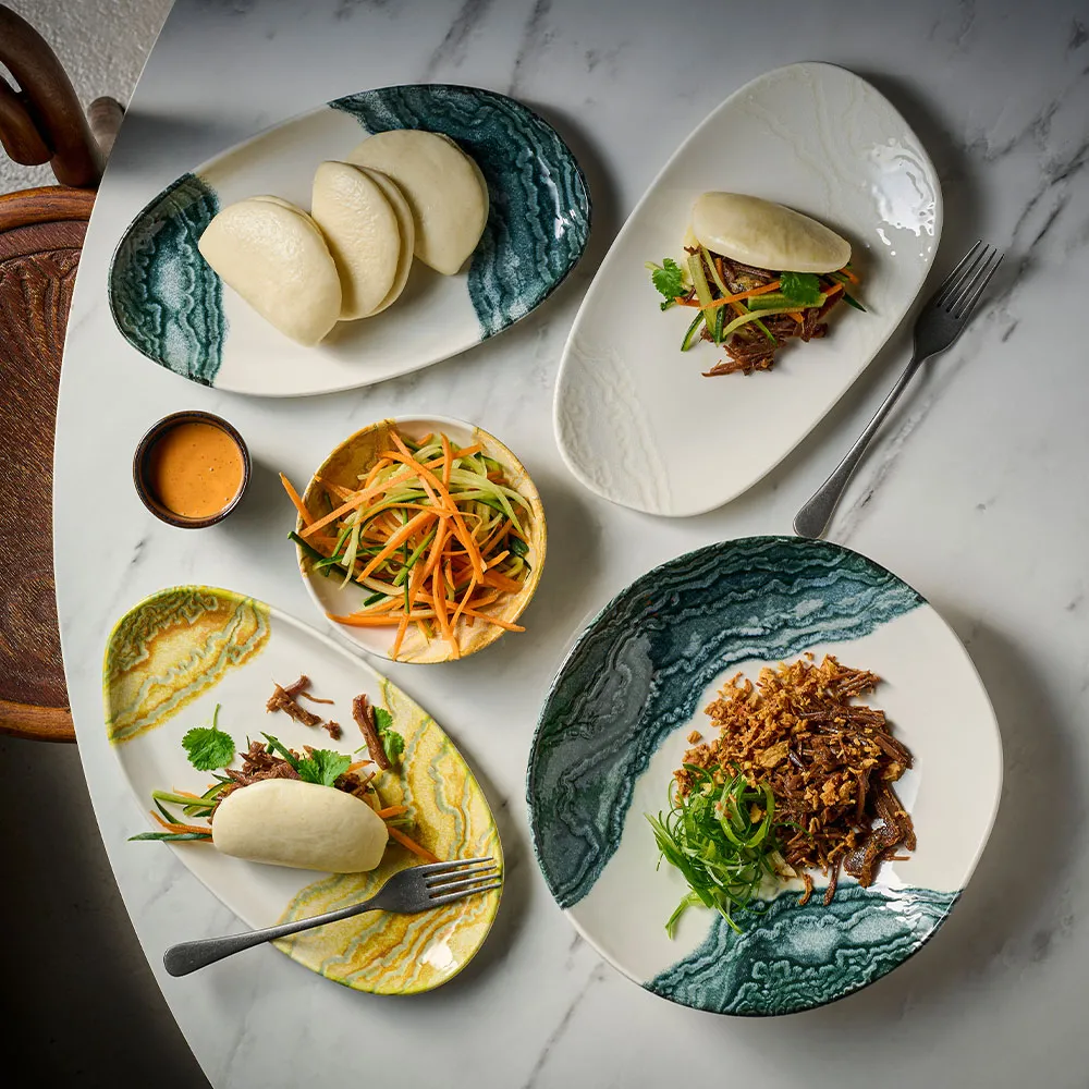 Churchill Inked Tide Black und Gold Mischung aus Coupe-Schalen und Opal-Platten, präsentiert ein asiatisch inspiriertes Tischarrangement mit Bao Buns, geraspeltem Gemüse und mehr